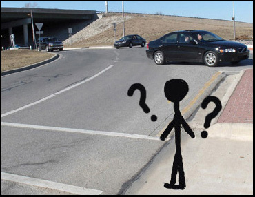 Photo shows a stick figure facing a crosswalk across two lanes.  Question marks are drawn around his head.  About 20 feet to our right, our street enters a roundabout (lanes go around a large circle).  Cars are coming from the other side of the circle and approaching the crosswalk.